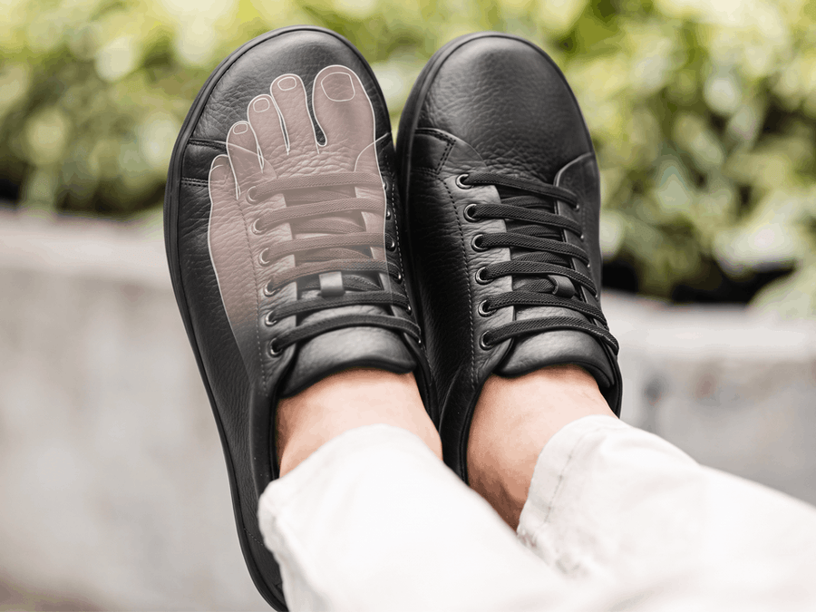 Black leather Birchbury sneakers with a translucent foot outline overlaid on one shoe, showing how the wide toe box gives toes room to spread naturally.
