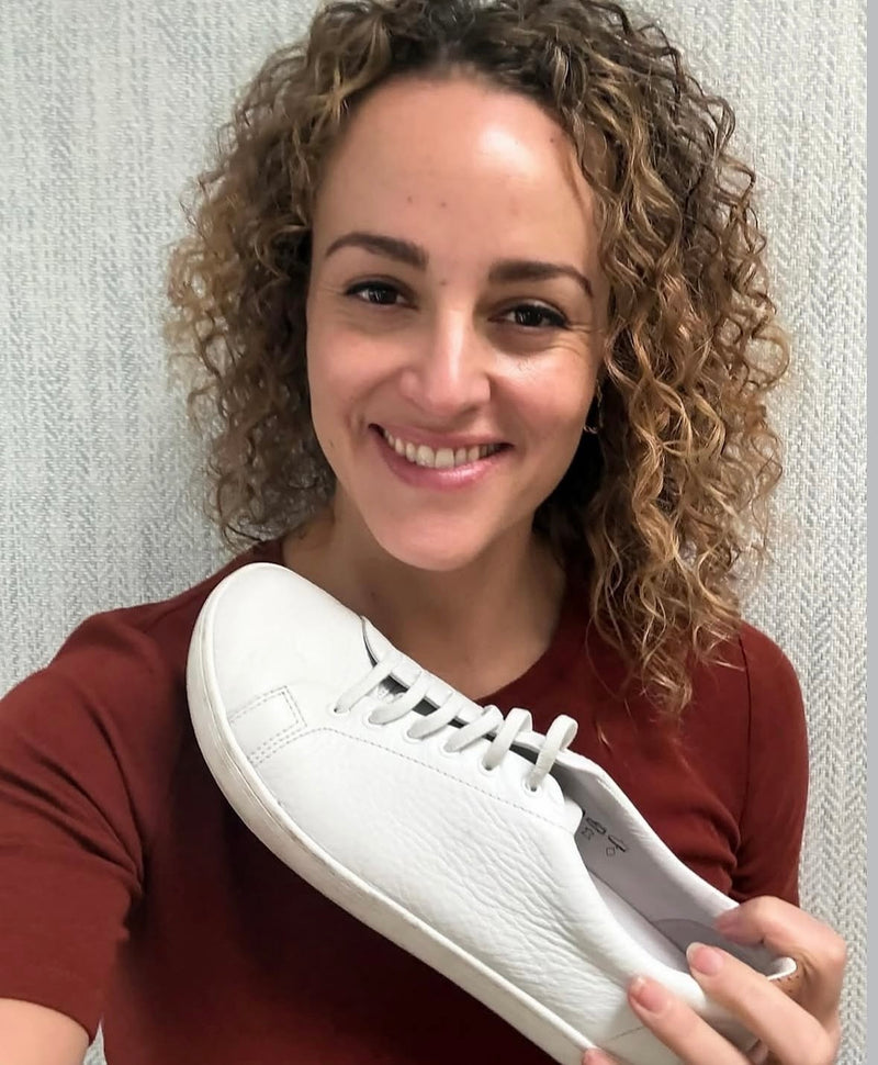 A smiling woman with curly hair holding up a white leather Birchbury sneaker with visible wear, standing against a textured gray wall.