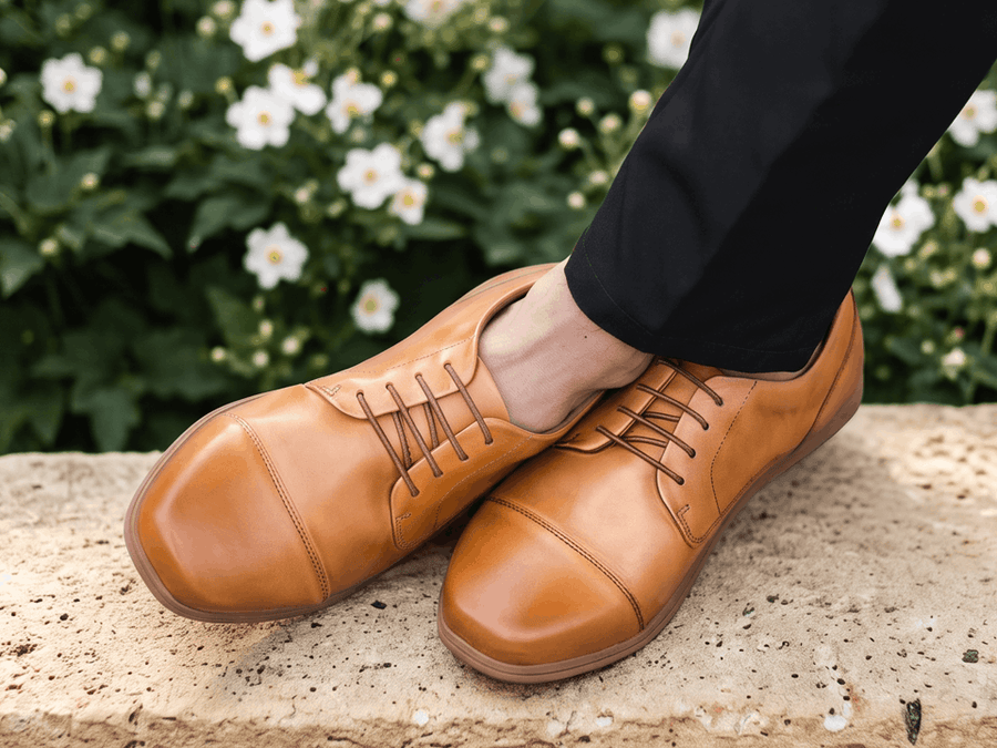 Overhead view of tan leather cap-toe Birchbury oxfords with crossed ankles, worn with black dress trousers on a stone-tiled sidewalk.