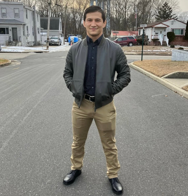 A man in a gray leather bomber jacket, navy polka dot shirt, and khaki pants standing on a suburban street, wearing black leather Birchbury oxfords, with houses and bare winter trees in the background