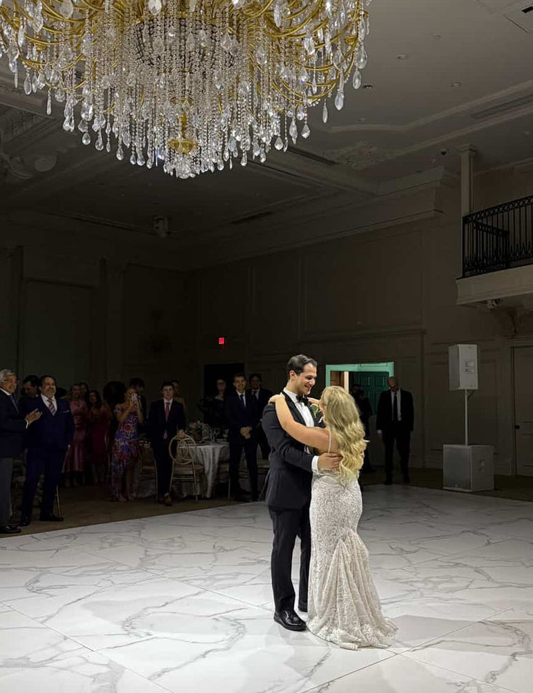 A groom in a black tuxedo and bride in a lace wedding gown share their first dance on a marble ballroom floor beneath an ornate crystal chandelier, surrounded by seated guests.