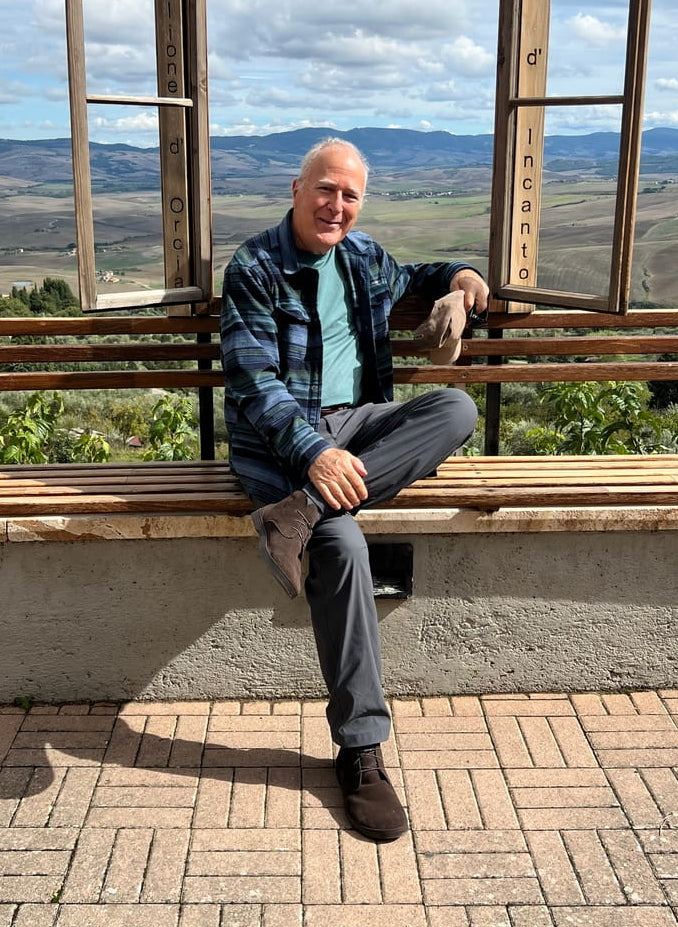 An older man in a blue plaid jacket smiling while seated on a bench in front of a decorative wooden window frame overlooking the Tuscan countryside, wearing brown suede Birchbury chukka boots.